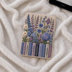 Peut inclure: Une housse de liseuse rectangulaire ornée d'une illustration détaillée de fleurs sauvages bleues et violettes, incluant des marguerites et des hortensias, disposées au-dessus d'une rangée de dos de livres verticaux aux motifs botaniques assortis.