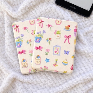 Peut inclure: Deux étuis Kindle crème avec un motif répété de nœuds roses, de fleurs et de petites illustrations. Les étuis sont rectangulaires avec des coins arrondis. Un appareil Kindle est visible en arrière-plan.