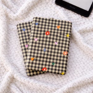 Peut inclure: Deux couvertures de livre à carreaux beiges et noirs avec des ornements colorés en forme de cœur, d'étoile et de fleur. Les couvertures sont posées sur une surface blanche texturée, avec un lecteur Kindle visible en arrière-plan.