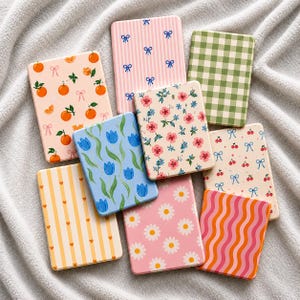May include: A selection of rectangular decorative items with diverse patterns. Designs include oranges, bows, flowers, cherries, stripes, and daisies. Colours include pink, green, orange, and blue. The items are arranged on a soft, grey surface.