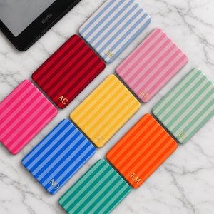 Pode incluir: Uma coleção de capas retangulares para leitores digitais com listras verticais em várias cores, incluindo vermelho, azul, amarelo, rosa, laranja e verde. Cada capa exibe um monograma dourado de duas letras na parte inferior, como AC, JM, KB, LK, EM, NO e LC.