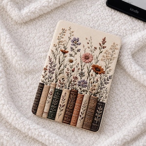 Op de afbeelding: Een beige Kindle-hoesje met een geborduurd ontwerp van kleurrijke wilde bloemen en een rij boeken in vintage-stijl. De hoes is rechthoekig en ligt op een witte, zachte deken. Een Kindle e-reader is zichtbaar op de achtergrond.