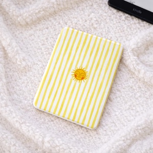 Puede incluir: Una funda Kindle a rayas amarillas y blancas con un diseño de sol bordado. La funda es rectangular y tiene un tacto suave y texturizado. El diseño del sol está en el centro y tiene una cara sonriente.