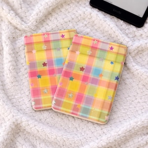 Peut inclure: Deux étuis rectangulaires avec un motif à carreaux coloré rose, jaune et bleu, ornés d'étoiles. Les étuis sont probablement pour un lecteur électronique ou une tablette. Le fond est une surface blanche texturée.