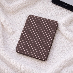 Könnte beinhalten: Eine braune Kindle-Hülle mit einem Muster aus kleinen, gleichmäßig verteilten rosa Punkten. Die Hülle ist rechteckig und scheint aus einem glatten Material zu bestehen. Im Hintergrund ist ein Kindle-E-Reader zu sehen.