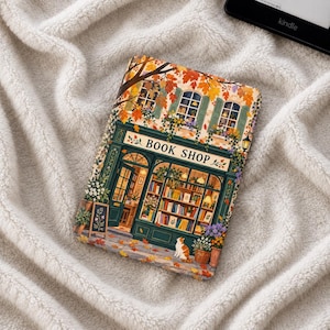 Puede incluir: Una funda rectangular para lector electrónico con una colorida ilustración de una librería con marcos verdes, grandes escaparates con estanterías de libros, hojas de otoño y un pequeño gato sentado en la acera frente a la entrada.