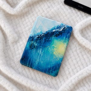 Peut inclure: Une housse de liseuse rectangulaire ornée d'une peinture abstraite aux nuances vibrantes de bleu marine profond, de bleu céruléen et de bleu ciel. Le motif présente un effet lumineux jaune circulaire et des coups de pinceau texturés.