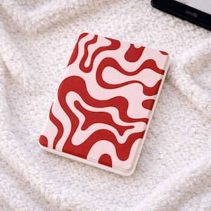 Peut inclure: Un étui Kindle rectangulaire avec un motif ondulé rouge et rose clair. L'étui a une bordure blanche et repose sur une couverture blanche et moelleuse. Un appareil Kindle est partiellement visible en arrière-plan.