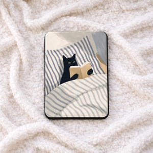 Peut inclure: Coque de téléphone noire rectangulaire avec une illustration d'un chat noir lisant un livre au lit. Le chat est sous une couverture à rayures, avec une palette de couleurs neutres.
