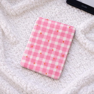 Peut inclure: Une housse de Kindle à motif vichy rose et blanc, ornée de petites étoiles orange. La housse rectangulaire est en tissu. En arrière-plan, une couverture blanche et moelleuse. Un Kindle est visible.