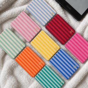Puede incluir: Una colección de fundas rectangulares para lectores electrónicos con rayas verticales en varios colores, incluyendo azul claro, rosa, rojo, verde, amarillo, naranja y azul oscuro. Cada funda muestra un pequeño monograma dorado en la esquina.
