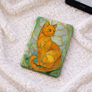 Peut inclure: Un étui rectangulaire avec un motif de style vitrail représentant un chat orange assis aux yeux jaunes. L'étui a une bordure turquoise et dorée et est posé sur un fond blanc et moelleux. Un lecteur Kindle est visible.