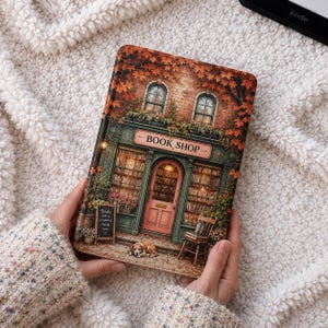 Può includere: Una custodia decorativa per Kindle con un'illustrazione dettagliata di una pittoresca libreria. La custodia presenta una facciata in mattoni, una tenda verde con il testo "BOOK SHOP" e foglie autunnali. Un piccolo cartello recita "Books are a universe inside you."
