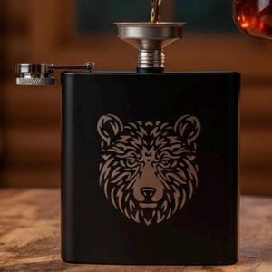 May include: A black flask with a bear design is being filled with amber liquid from a glass bottle. The flask is on a wooden table, with a fireplace and books in the background. The flask has a hinged cap.