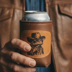 Könnte beinhalten: Ein brauner Leder-Dosenkühler mit einem Cowboy-Frosch-Motiv. Der Kühler wird von einer Person gehalten, die eine braune Lederjacke und einen Cowboyhut trägt. Die Dose im Inneren ist silberfarben.