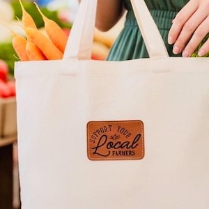 Op de afbeelding: Een crèmekleurige draagtas met een bruin label met de tekst "Support Your Local Farmers". De tas bevat wortelen en groene groenten. De foto is genomen op een boerenmarkt, met verse producten op de achtergrond.