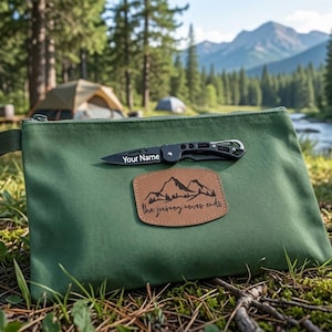 May include: A rectangular olive green canvas pouch featuring a tan leather patch with a mountain graphic and the text 'the journey never ends'. A black folding pocket knife with the text 'Your Name' printed on the blade rests on the pouch.