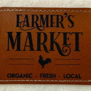 May include: A rectangular, brown leather patch with the words "FARMER'S MARKET" in black, stylized font. Below the text is a rooster silhouette and the words "ORGANIC - FRESH - LOCAL". The patch has a stitched border.