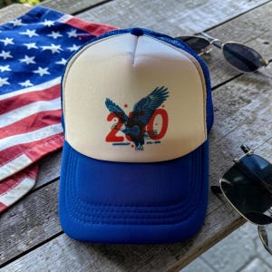 May include: A blue and white trucker hat with a blue brim and a white front panel. The front panel features a graphic of a blue eagle and the number 20, with the text "Anniversary 1776-2026".