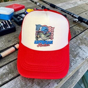 May include: A red and white trucker hat with a red brim and mesh back. The front panel features a graphic of a fish wearing sunglasses and an American flag, with the text "American Hooker".