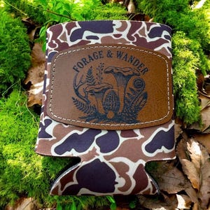 Op de afbeelding: Een drankhouder met camouflagepatroon en een bruin leren label. Het label toont de woorden "FORAGE & WANDER" en een afbeelding van paddenstoelen en bladeren. De houder is ontworpen om drankjes koel te houden.