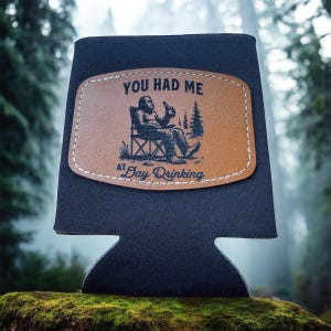 Könnte beinhalten: Schwarzer Getränkehalter mit einem braunen Lederaufnäher. Der Aufnäher zeigt eine Bigfoot-Illustration, die in einem Stuhl sitzt und eine Flasche hält, mit dem Text "YOU HAD ME AT Day Drinking". Entwickelt, um Getränke kühl zu halten.