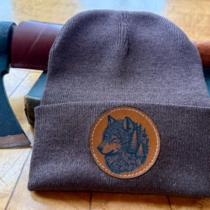 May include: A dark brown knit beanie featuring a circular tan leather patch on the front. The patch displays a detailed dark blue illustration of a wolf head surrounded by pine trees. A dark metal hatchet with a wooden handle is positioned behind.