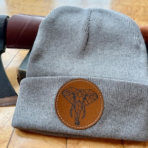 May include: A gray knit beanie with a brown leather patch featuring a geometric elephant design. An axe with a black head and wooden handle rests next to the hat, along with a book.