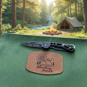Puede incluir: Un estuche de lona verde con cremallera y un parche de cuero tostado con un gráfico de sol, montañas y árboles con el texto Adventure Awaits. Una navaja plegable negra con una hoja plateada grabada con el texto Your Name descansa encima.