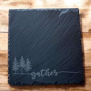 May include: A square, dark grey slate serving board with a textured surface. The board features an etched design of three stylized pine trees and the word "gather" in a cursive font. The board is set against a wooden background.
