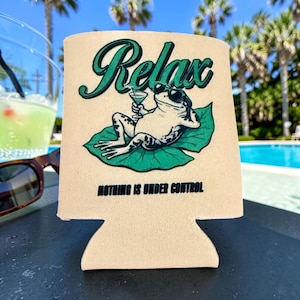 May include: A beige foam can cooler featuring a graphic of a frog wearing sunglasses, lounging on a green lily pad while holding a cocktail. The text Relax is printed in green above the frog, and Nothing is under control is printed below.