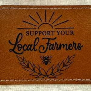 May include: A rectangular, brown leather patch with the text "SUPPORT YOUR Local Farmers" in black script. The design includes a sunburst, a bee, and a laurel wreath. The patch has a stitched border.