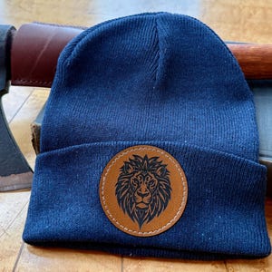 May include: A navy blue knit beanie with a brown leather patch featuring a lion's head. An axe and a book are in the background. The hat is a stylish accessory for outdoor activities or casual wear.