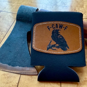 May include: A black beverage holder with a brown leather patch featuring a raven illustration and the text "F-CAW-F". The holder is next to a blue axe head with a wooden handle.