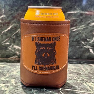 Könnte beinhalten: Ein brauner Leder-Dosenkühler mit einem Waschbär-Motiv und dem Text "IF I SHENAN ONCE I'LL SHENANIGAN". Eine gelbe Dose alkoholfreien Getränks befindet sich im Inneren. Der Kühler ist zum Kühlen von Getränken konzipiert.