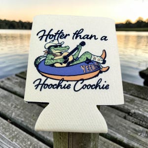 K&ouml;nnte beinhalten: Ein wei&szlig;er Dosenk&uuml;hler aus Schaumstoff mit einer Grafik eines gr&uuml;nen Alligators, der einen Cowboyhut und eine Sonnenbrille tr&auml;gt und in einem blau-orangen Schwimmreifen Gitarre spielt, mit dem schwarzen Schriftzug Hotter than a HOOCHIE COOCHIE.