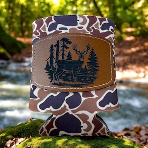 Puede incluir: Un enfriador de latas con estampado de camuflaje con un parche de cuero marrón con la silueta de un ciervo en una escena forestal. El enfriador tiene un patrón marrón, morado y blanco.