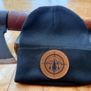 May include: A navy blue knit beanie with a brown leather patch featuring a compass design with mountains and trees. An axe with a wooden handle and a leather sheath is visible in the background, along with a book.