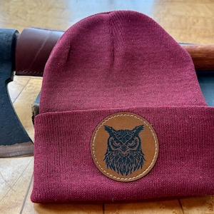 May include: A claret-coloured knitted beanie hat with a brown leather owl patch. An axe with a leather sheath and a wooden handle rests behind the hat, along with a vintage book.