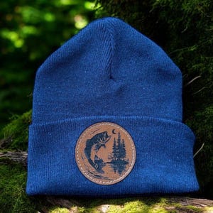 Puede incluir: Un gorro azul marino con un parche circular marrón que representa un pez saltando, árboles y una luna creciente. El parche está cosido en el borde doblado. El fondo es verde borroso.