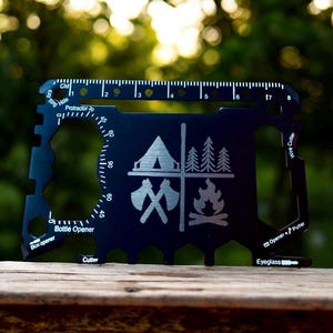 Può includere: Multi-tool nero con grafica a tema campeggio, tra cui una tenda, alberi, asce e un falò. Dispone di un righello con misure in cm, un goniometro e varie funzioni di utensili come un apribottiglie e un tagliafili.
