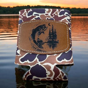 May include: Camouflage-patterned can cooler with a brown leather patch. The patch features a leaping fish graphic with a crescent moon and trees. The cooler is designed to keep beverages cold and is ideal for fishing enthusiasts.