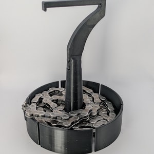 May include: A black bicycle chain tool with a handle and a hook, holding a silver bicycle chain. The tool is designed to assist with chain maintenance and repair. The chain is coiled within a black circular base.