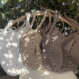 May include: Four handmade crochet tote bags in white, beige, and brown. Each bag has a unique texture and design, with braided handles and some featuring decorative tassels. The bags are displayed on a wicker chair.