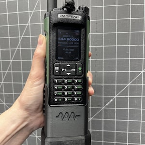 May include: A handheld Baofeng radio in olive green and black, held by a hand. The radio has a screen displaying "DANSVILLE 444.60000" and other data. It features a keypad with numbers and function keys, and a prominent antenna.