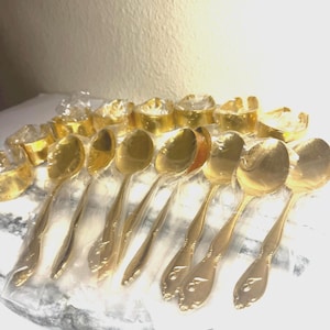 Puede incluir: Una colección de cucharas de metal de color dorado y servilleteros a juego, cada uno envuelto individualmente en plástico transparente. Las cucharas presentan un acabado liso y reflectante con un detalle en relieve en el mango.