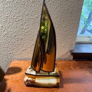 May include: A decorative sailboat sculpture featuring two tall, amber-colored glass sails with dark metal edges. The boat hull is also crafted from amber glass, and the entire structure is mounted on a rectangular, polished white and tan stone base.