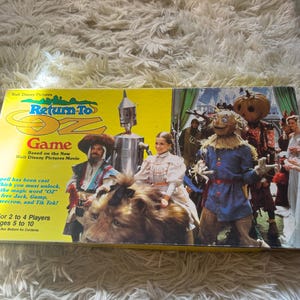 May include: A rectangular Return to Oz board game box featuring a yellow background with the title in blue and red text. The box displays characters from the movie including a tin robot, a lion, a scarecrow, and a pumpkin-headed figure.