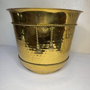 Puede incluir: Una maceta de latón pulido con una textura martillada en la superficie exterior. El recipiente está decorado con dos bandas horizontales estilo cuerda y dos borlas colgantes en la parte frontal, con un borde ancho y acampanado en la parte superior.