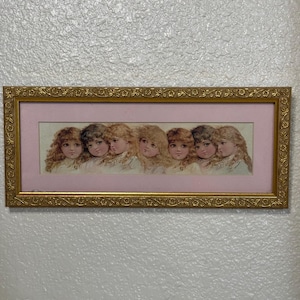 Puede incluir: Un cuadro rectangular que presenta una impresión horizontal de siete niños pequeños con cabello rizado dispuestos en fila. La impresión está colocada dentro de un paspartú rosa claro y encerrada en un marco dorado texturizado con detalles florales.